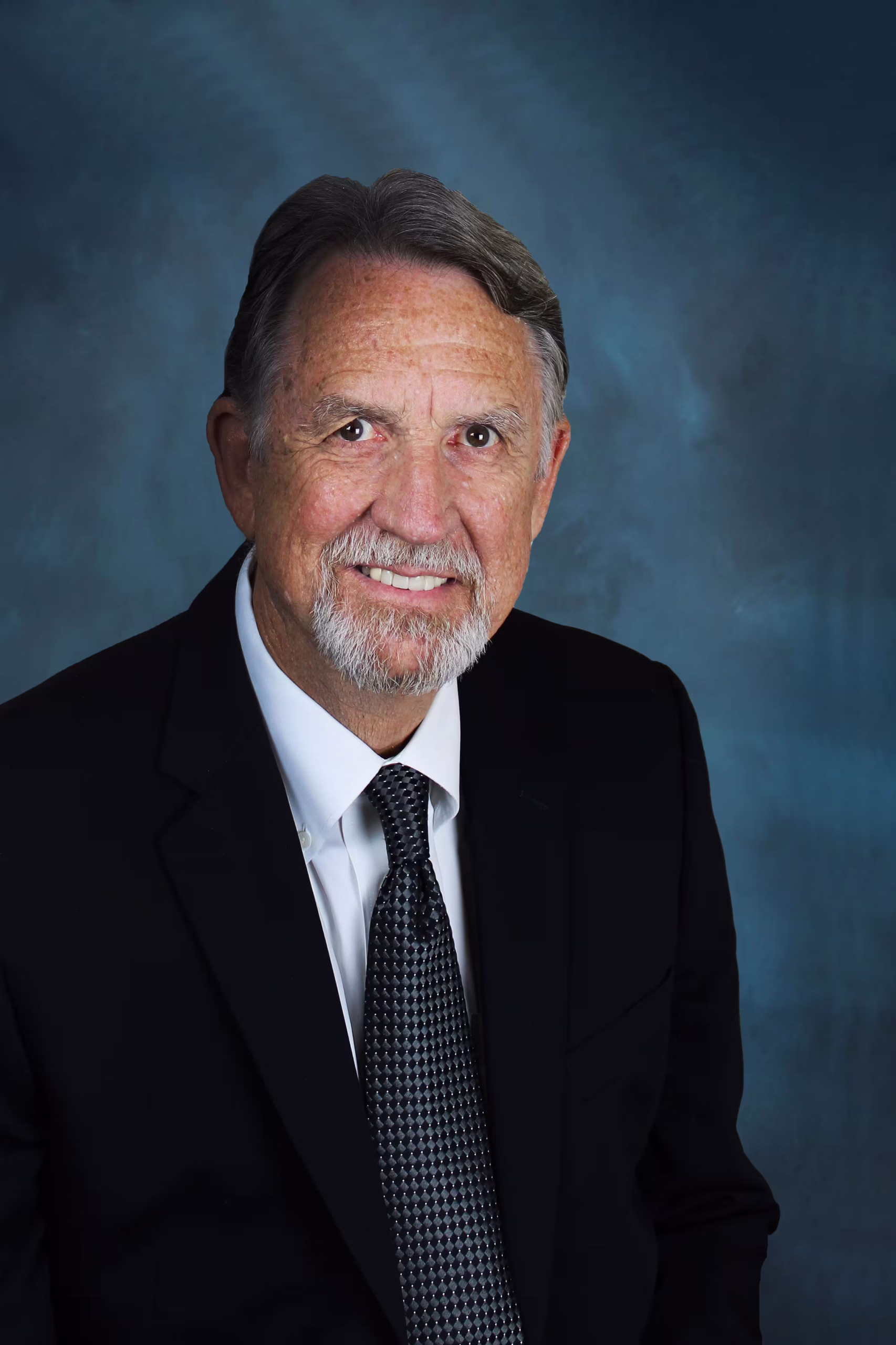 Official portrait of Councilman Steele, a smiling older gentleman with gray hair and a white goatee wearing a black suit, white dress shirt, and patterned charcoal tie against a blue backdrop