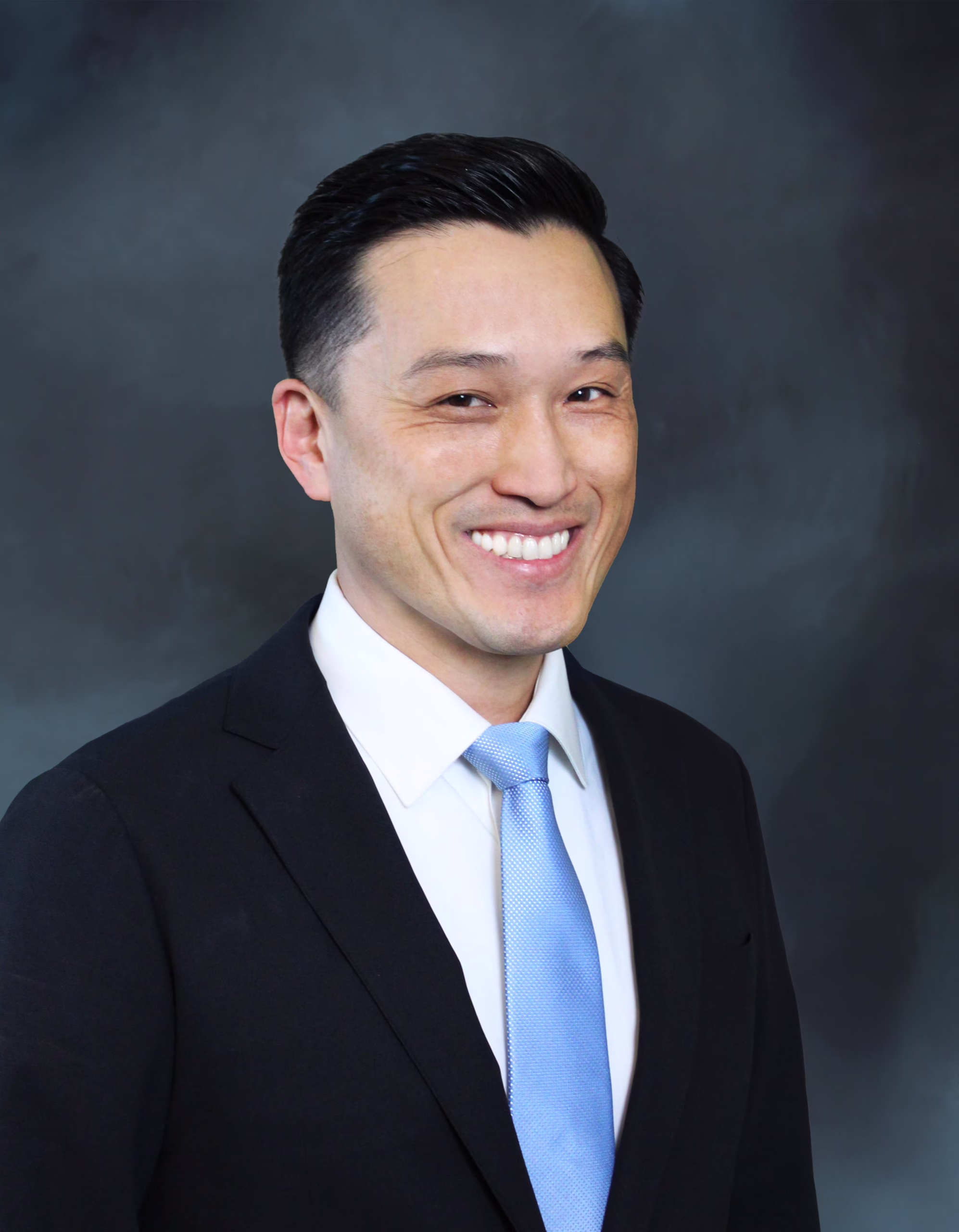 Professional headshot of a smiling businessman with short dark hair wearing a navy suit, white dress shirt, and light blue tie against a gray studio backdrop